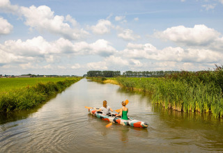 Twee mensen peddelen in een kleurrijke kajak door een rustige sloot tussen groene weilanden in Gelderland, Nederland.
