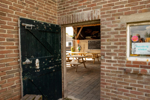 Eingang mit offener Holztür, Blick auf Sitzbänke und Tafel bei Feather Down Hoeve de Betuwe, Niederlande.