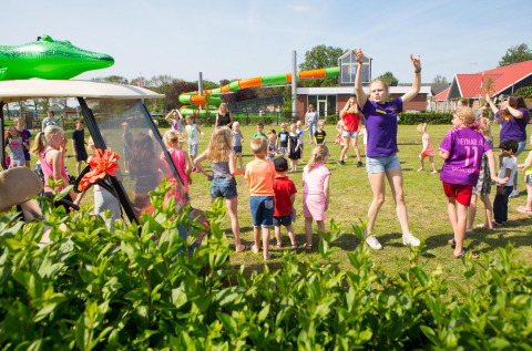 Kinderen en volwassenen spelen samen op een campingveld met safaritenten en glijbanen op de achtergrond.
