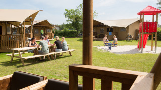 Famiglie si rilassano davanti a tende safari con parco giochi al Camping 't Veld nei Paesi Bassi.