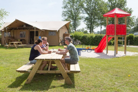 Three adults sit at a picnic table in front of a safari tent at Camping 't Veld with a playground nearby.