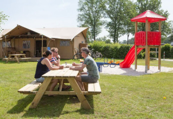 Three adults sit at a picnic table in front of a safari tent at Camping 't Veld with a playground nearby.
