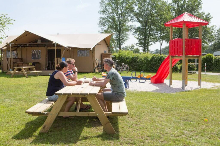 Three adults sit at a picnic table in front of a safari tent at Camping 't Veld with a playground nearby.