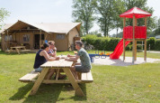 Three adults sit at a picnic table in front of a safari tent at Camping 't Veld with a playground nearby.
