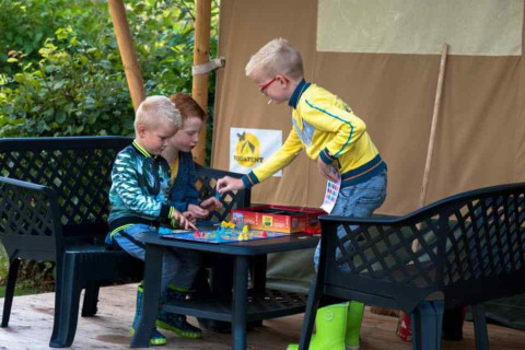 Tres niños juegan a un juego de mesa fuera de una tienda safari en Camping 't Veld, Países Bajos, rodeados de naturaleza.