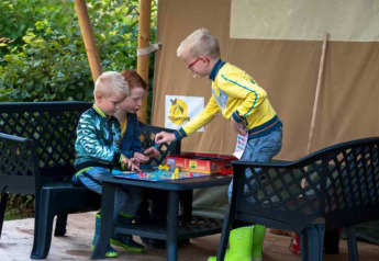 Tres niños juegan a un juego de mesa fuera de una tienda safari en Camping 't Veld, Países Bajos, rodeados de naturaleza.