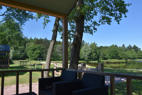 Vista desde la Beach House lodge en Camping 't Veld, Países Bajos, con terraza, lago y vegetación.