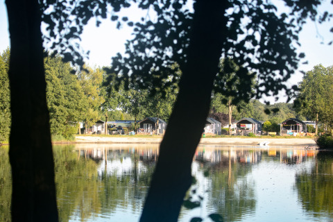 Udsigt over søen til strandhytter på Camping 't Veld, Nederlandene, omgivet af træer og natur.