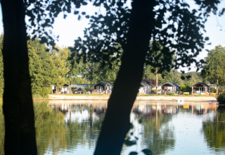 View across a lake towards beach houses at Camping 't Veld, Netherlands, nestled among trees and nature.