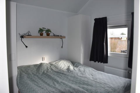 Pequeño dormitorio con cama doble, estante con plantas, ventana y cortina negra en Beach house, Camping 't Veld.