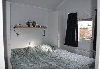 Pequeño dormitorio con cama doble, estante con plantas, ventana y cortina negra en Beach house, Camping 't Veld.