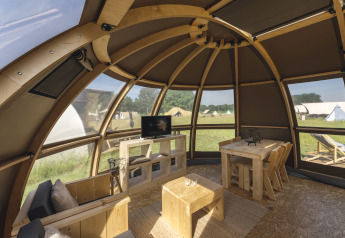 Intérieur d’un dôme de glamping de luxe avec meubles en bois, grandes fenêtres et vue sur les tentes.