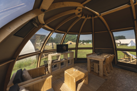 Innenraum einer luxuriösen Glamping-Kuppel mit Holzmöbeln, großen Fenstern und Blick auf Zelte draußen.
