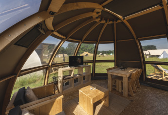 Interieur van een luxe glampingkoepel met houten meubels, grote ramen en uitzicht op tenten buiten.