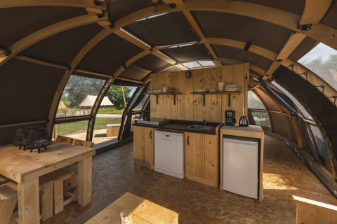 Vista interior de una tienda glamping con cocina pequeña, mesa de madera y grandes ventanas hacia la naturaleza.