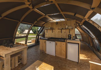 Binnenaanzicht van een glampingtent met kleine keuken, houten tafel en grote ramen met zicht op de natuur.