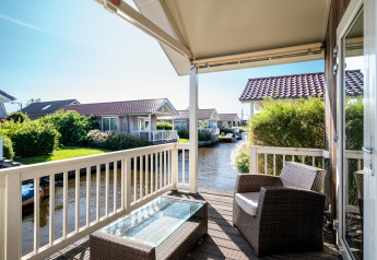 Zicht vanaf het terras van een lodge met wicker stoelen, glazen tafel en huisjes aan een kanaal.