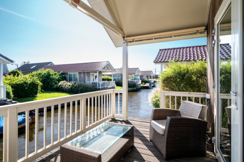 View from a lodge terrace with wicker chairs, a glass table, and canal-side cottages in the sunlight.