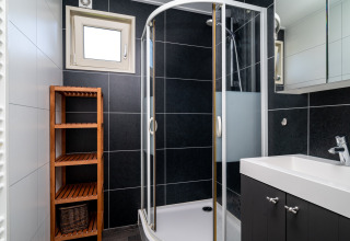 Baño moderno en lodge con ducha de cristal, lavabo blanco, estante de madera y azulejos negros en la pared.