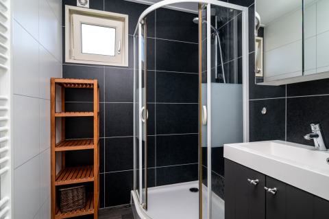 Moderne lodgebadkamer met glazen douchecabine, witte wastafel, houten rek en zwarte tegelwanden.