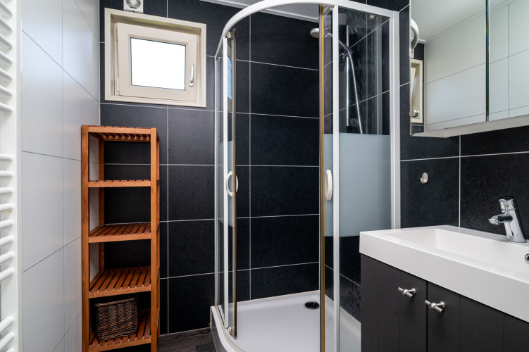Baño moderno en lodge con ducha de cristal, lavabo blanco, estante de madera y azulejos negros en la pared.