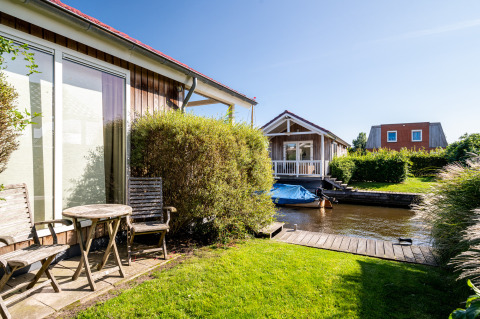 View of a lodge with wooden furniture, garden, and a small canal with boats under a clear blue sky.