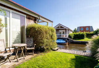 Udsigt over en lodge med træmøbler, have og en lille kanal med både under en klar blå himmel.