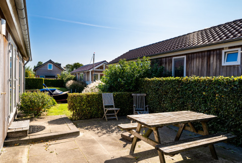 Buitenterras bij een lodge met houten picknicktafel, stoelen, hagen en zonnige blauwe lucht.