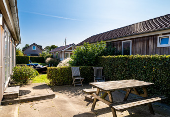 Buitenterras aan een lodge met houten tafel, stoelen, groene hagen en een zonnige lucht.