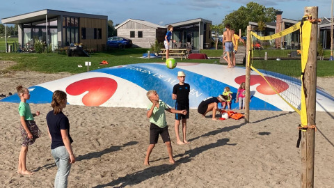 Niños juegan voleibol en una cancha de arena frente a cabañas glamping, otros saltan en un cojín inflable grande.