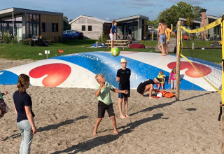 Niños juegan voleibol en una cancha de arena frente a cabañas glamping, otros saltan en un cojín inflable grande.
