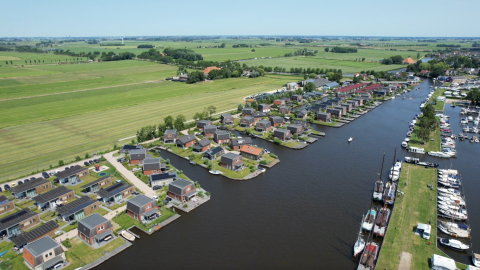 Vista aérea de alojamiento glamping junto a un río con modernas cabañas, barcos y campos verdes.
