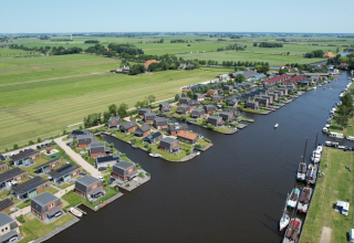 Vista aérea de alojamiento glamping junto a un río con modernas cabañas, barcos y campos verdes.