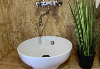 Moderne vask med hvid håndvask, kromarmatur og grøn plante på træbord i luksustelt ved La Draille.