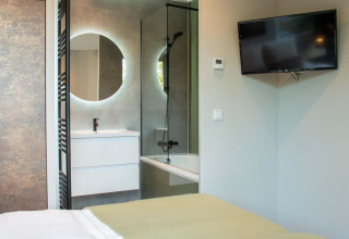 Dormitorio moderno con televisor de pared y vista al baño con espejo redondo y bañera en Veluwehoeve Wellness Lodge.