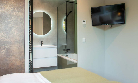 Moderne slaapkamer met tv aan de muur, zicht op badkamer met ronde spiegel en bad bij Veluwehoeve Wellness Lodge.