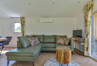 Woonkamer van een luxe lodge met groene zetel, ronde houten tafel, tv en grote ramen bij Drentse Weelde, Nederland.