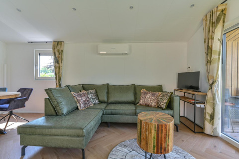 Woonkamer van een luxe lodge met groene zetel, ronde houten tafel, tv en grote ramen bij Drentse Weelde, Nederland.