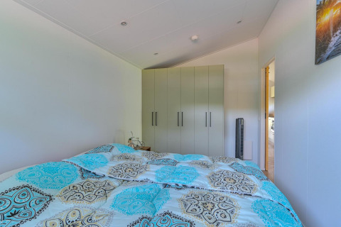 Dormitorio con edredón estampado, armario y vista a la cocina en la Luxury lodge de Drentse Weelde.