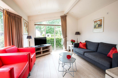 Lichte zithoek met rode zetels, grijze sofa en groot raam in een vakantiehuis bij EuroParcs Noordwijkse Duinen.