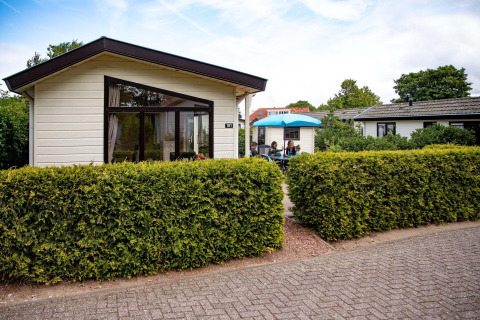 Feriehytte ved EuroParcs Noordwijkse Duinen i Holland, med terrasse, hæk og folk under en blå parasol.