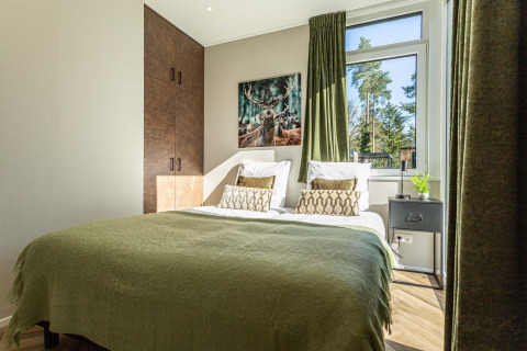 Acogedor dormitorio de lodge con cama doble, colcha verde, vista al bosque y cuadro de ciervo en la pared.