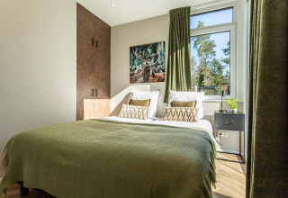 Acogedor dormitorio de lodge con cama doble, colcha verde, vista al bosque y cuadro de ciervo en la pared.