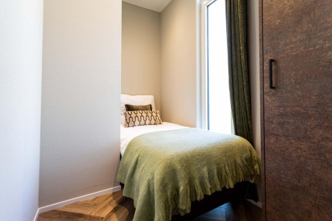 Dormitorio pequeño y moderno en una cabaña, con cama individual, manta verde y gran ventanal.