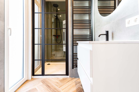 Baño moderno en cabaña con puerta de ducha de vidrio, grifería negra y tocador blanco sobre suelo de madera.