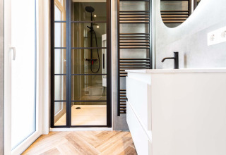 Baño moderno en cabaña con puerta de ducha de vidrio, grifería negra y tocador blanco sobre suelo de madera.