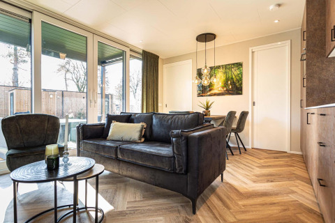 Lichte, moderne woonkamer in een lodge met grote ramen, parketvloer en eigentijds zwart meubilair.
