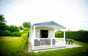 Lille hus med overdækket veranda og bænk, Forest Lodge i Holiday Park Duinhoeve, Nederlandene, omgivet af grønt.