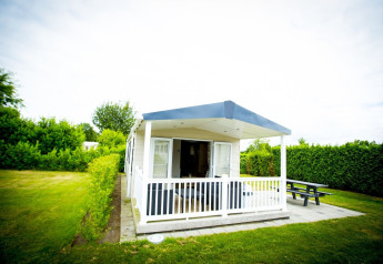 Lille hus med overdækket veranda og bænk, Forest Lodge i Holiday Park Duinhoeve, Nederlandene, omgivet af grønt.