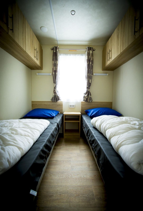 Kleine slaapkamer met twee eenpersoonsbedden, raam en houten kasten in Forest Lodge, Holiday Park Duinhoeve.